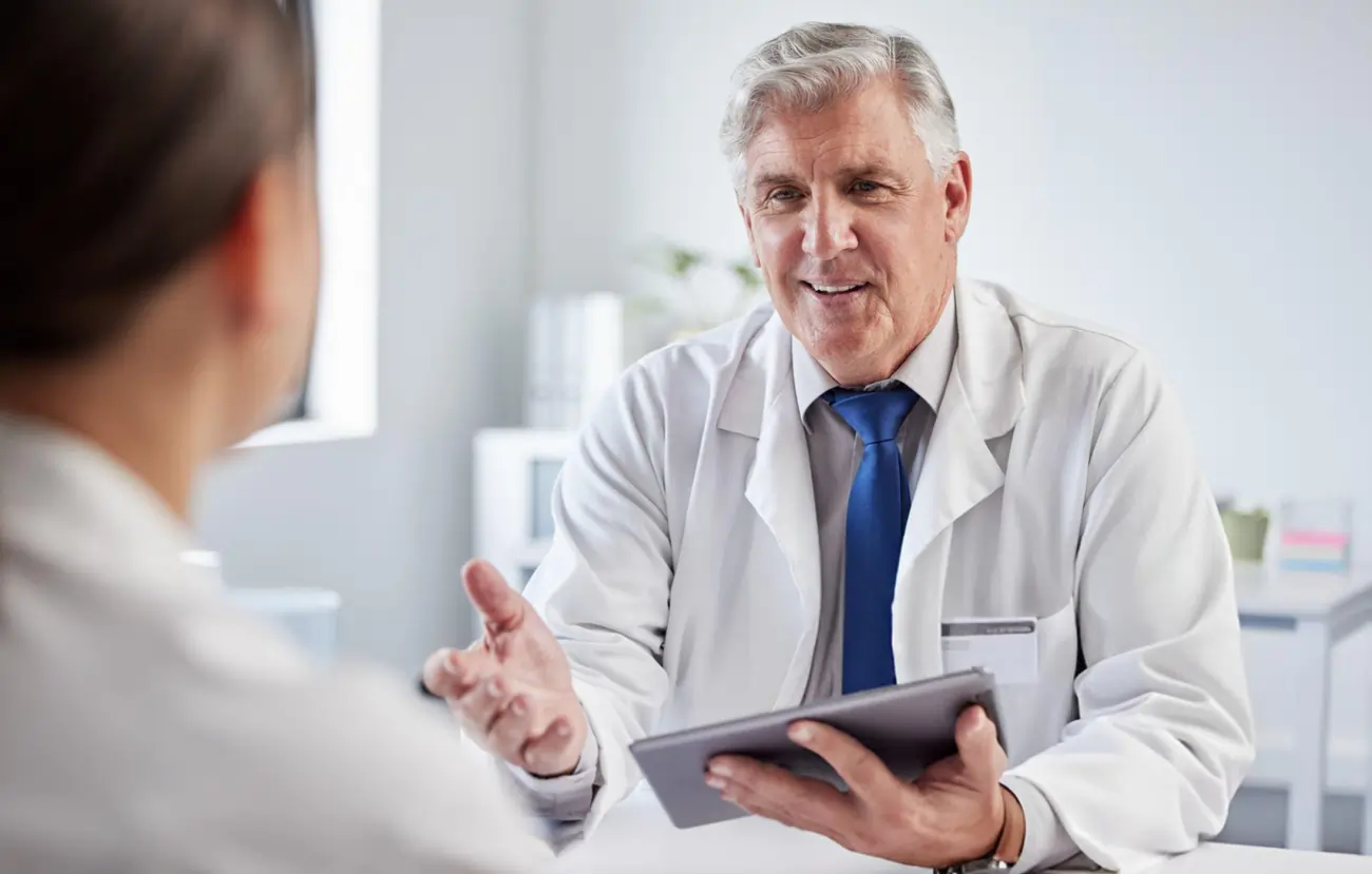 A compassionate, experienced male doctor in a white coat explains stem cell therapy options to a patient using a tablet in a clinical setting in Waco, Texas.