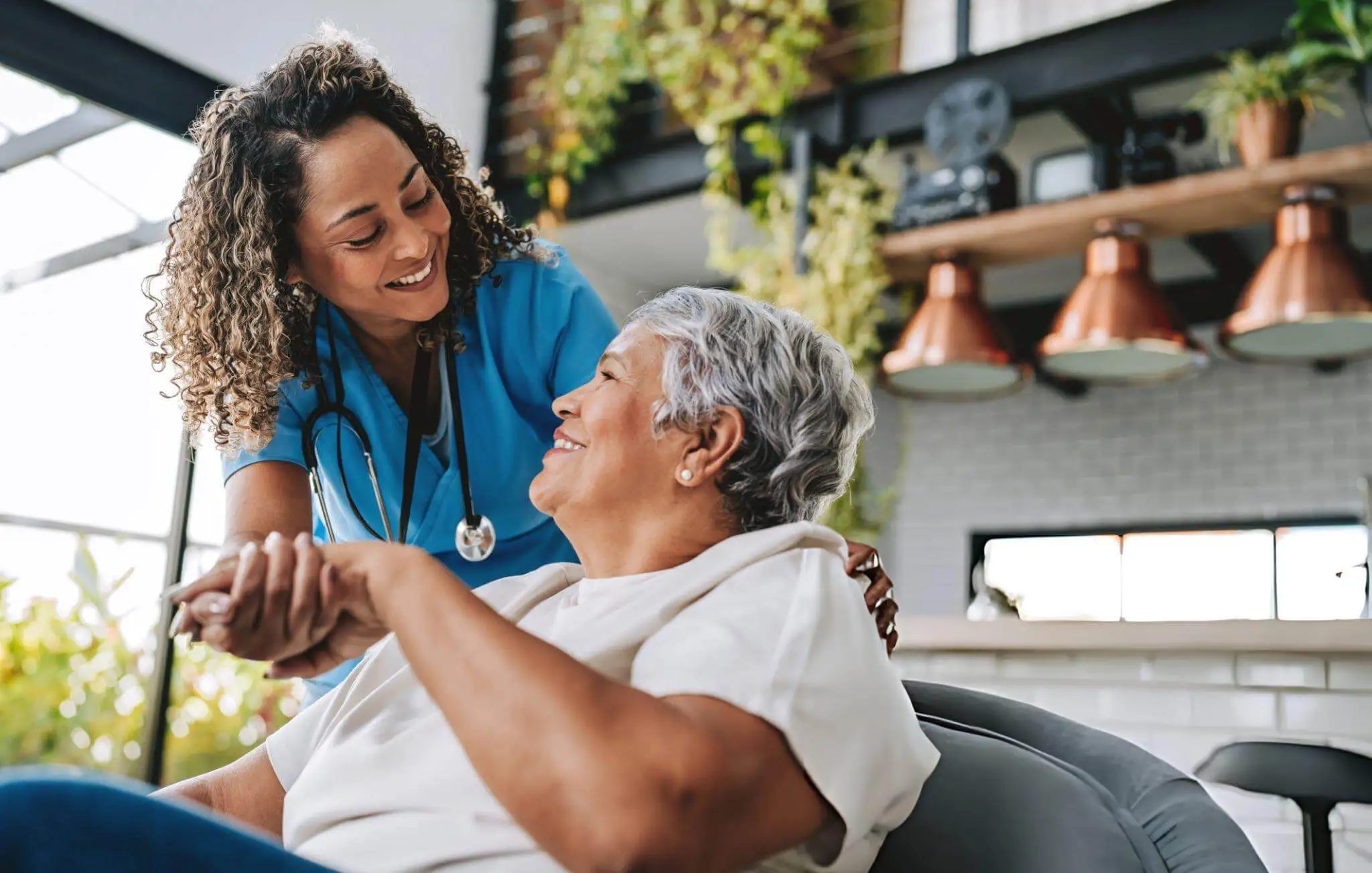 A smiling healthcare provider in blue scrubs and a stethoscope holding the hand of an elderly female patient in a bright, modern clinic in Winston-Salem