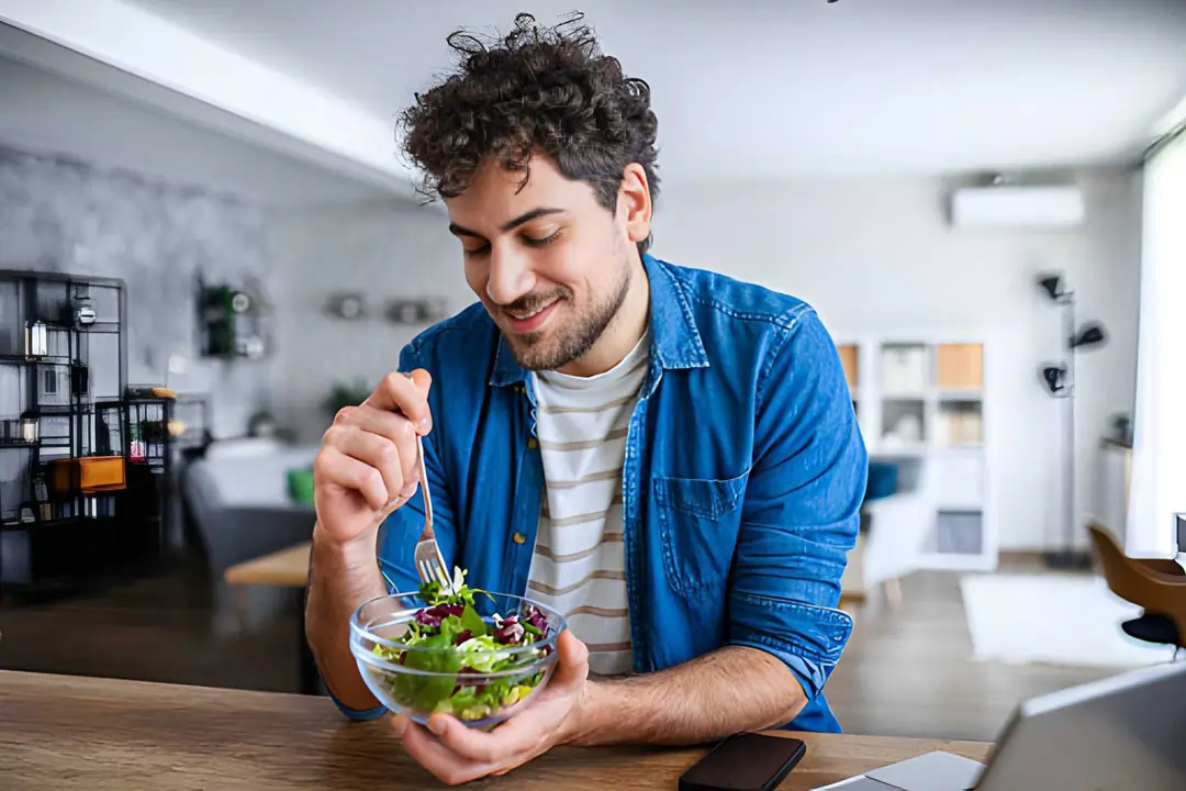 A young man smiling while eating a fresh, nutritious green salad in a bright home setting, representing the healthy diet and lifestyle changes recommended by R3 Stem Cell providers in Cape Coral, Florida