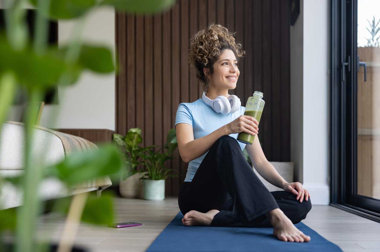 A young woman smiling and holding a healthy green smoothie while resting on a yoga mat, illustrating recovery after regenerative cell therapy.