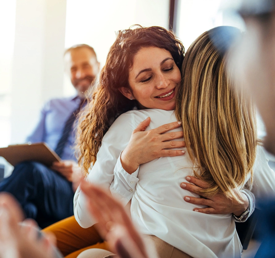 A smiling woman with curly hair warmly embracing another woman in a professional clinical setting, while a male healthcare provider watches supportively in the background.