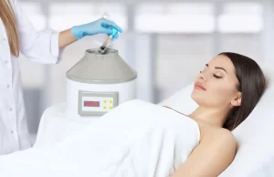 Woman receiving a facial cryotherapy or skin rejuvenation treatment at a beauty clinic while a technician handles a medical aesthetic device.