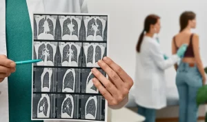 A medical professional in a green tie holds a sheet of lung CT scans, pointing a pen at specific results. In the background, a doctor in a white lab coat examines a patient.