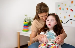 A smiling woman supporting a young girl from behind, helping her hands hold and play with a large, brightly colored swirl toy in a playroom.