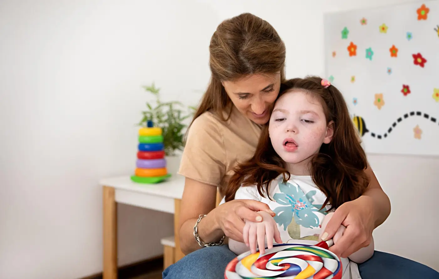 A smiling woman supporting a young girl from behind, helping her hands hold and play with a large, brightly colored swirl toy in a playroom.