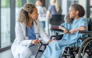 A female doctor in a white lab coat kneels to speak caringly with a smiling young girl sitting in a wheelchair in a bright hospital hallway.