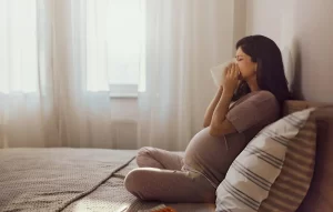 Pregnant woman sitting on bed blowing her nose into a tissue, experiencing cold or flu-like symptoms during pregnancy