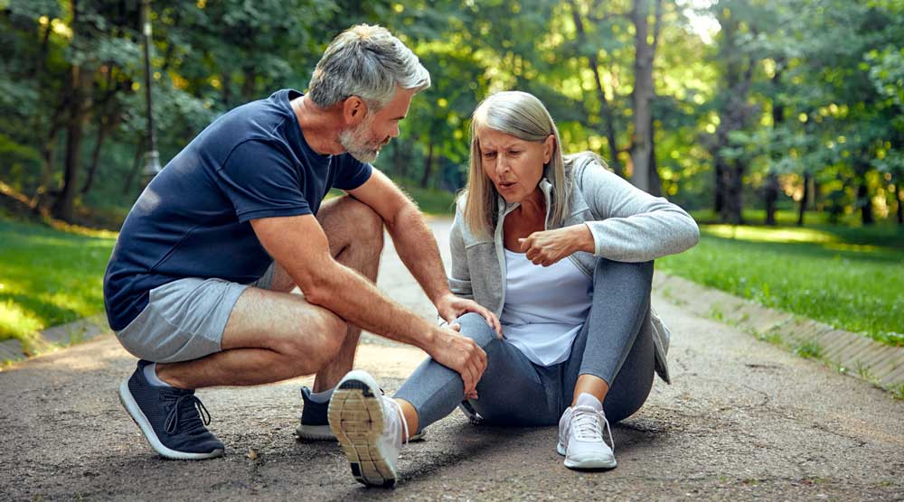 A senior woman sitting on a park path holding her injured knee in pain, while a supportive older man kneels down to comfort and check on her during a run.