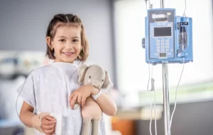 A young girl in a hospital gown smiles while holding a stuffed elephant toy, with an IV line in her hand and a medical infusion pump on a pole beside her.