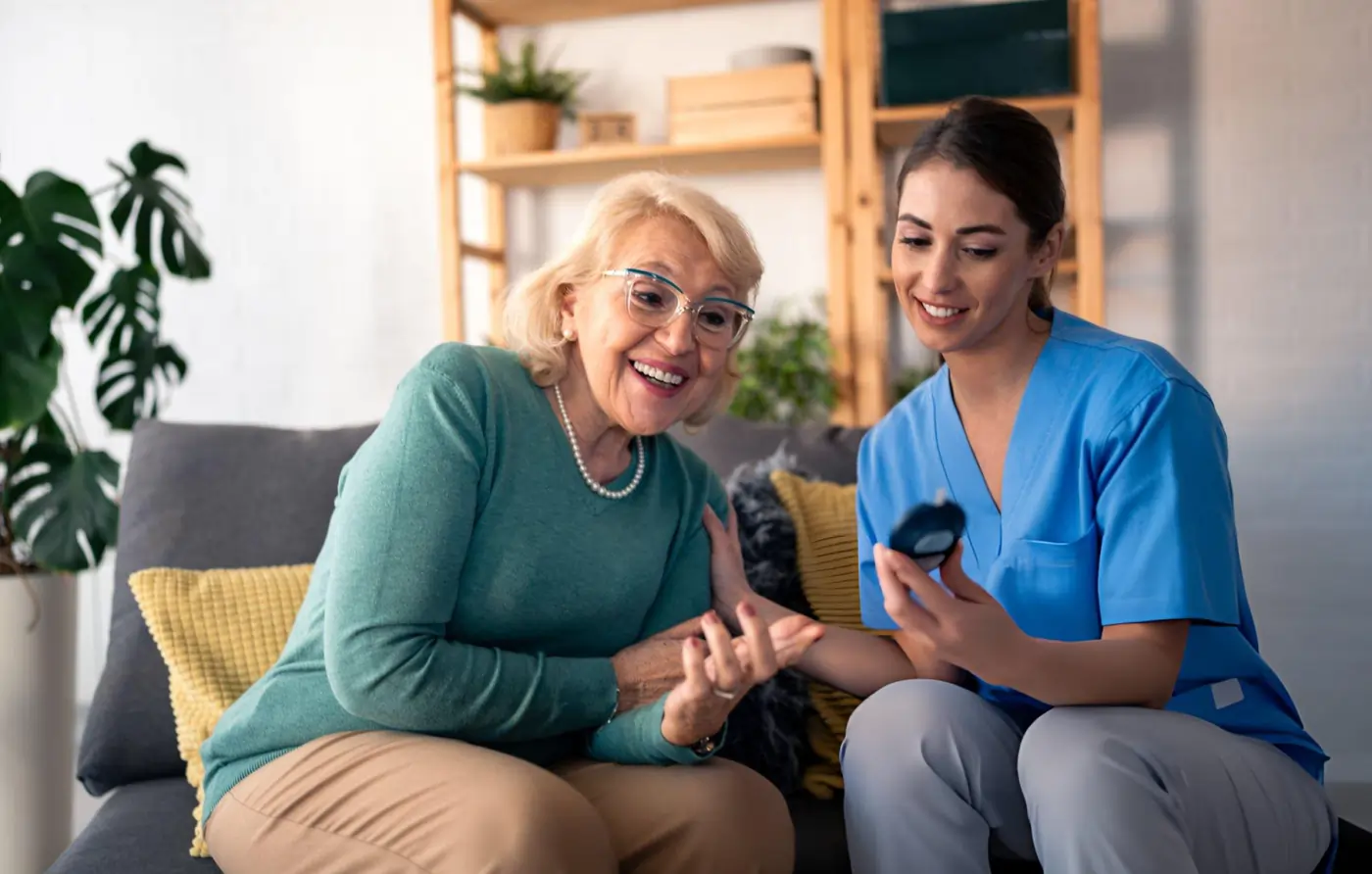 A nurse in blue medical scrubs sitting on a couch next to a smiling elderly female patient, showing her the blood glucose reading on a portable digital glucometer during a home care visit.