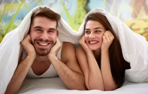 Smiling young couple lying under a blanket on a bed, looking happy and relaxed together in a cozy indoor setting.