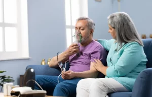 An elderly man sitting on a sofa and using a compressor nebulizer mask for respiratory therapy while a supportive caregiver sits beside him.