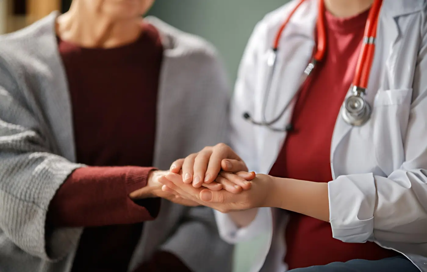 Close-up of a healthcare professional in a white coat and red stethoscope holding an elderly patient's hand during a consultation for preventative cell therapy, illustrating compassionate patient education.