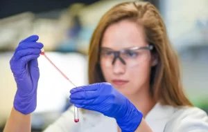 A medical laboratory researcher wearing blue gloves and safety glasses using a pipette to transfer a red liquid sample into a test tube for stem cell analysis.
