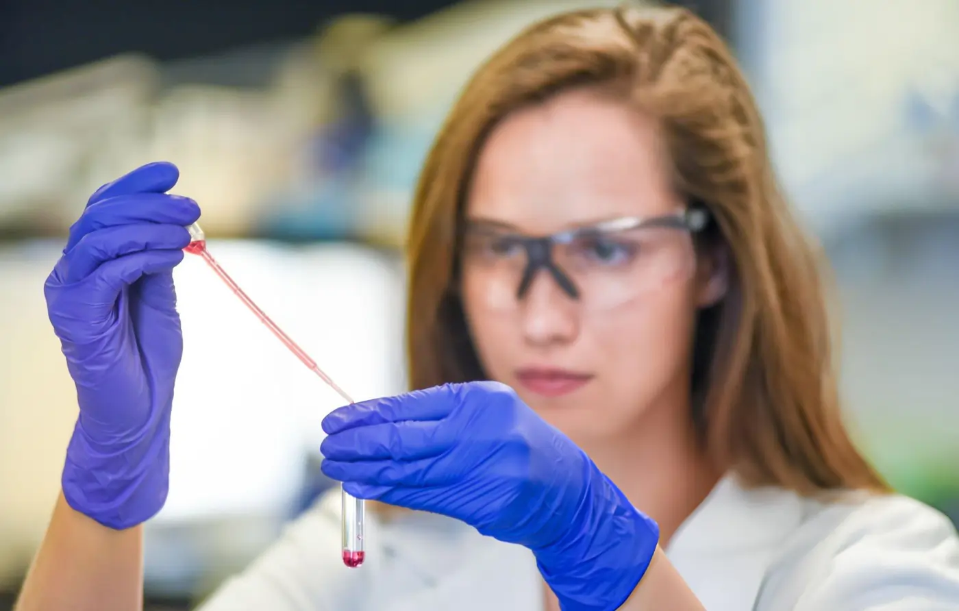 A medical laboratory researcher wearing blue gloves and safety glasses using a pipette to transfer a red liquid sample into a test tube for stem cell analysis.