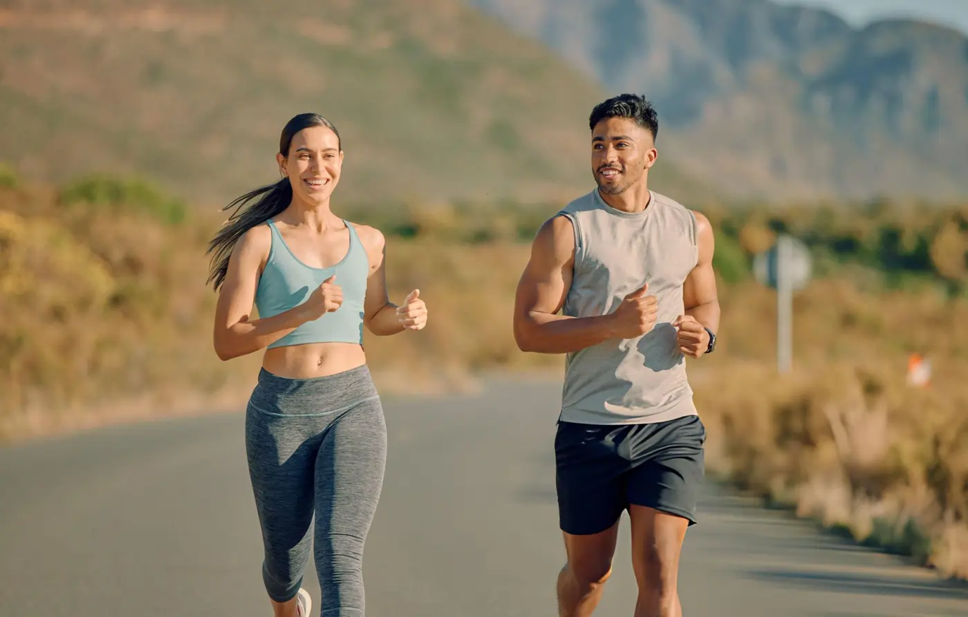 A fit young man and woman smiling and running together on an open outdoor road, representing healthy joint function and successful recovery from sports injuries or chronic pain.