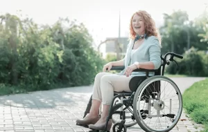 A smiling, curly-haired middle-aged woman sitting in a wheelchair on an outdoor paved path, reflecting positive functional improvements from multiple sclerosis treatment.