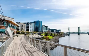 Waterfront boardwalk along the Columbia River in Vancouver, Washington, with modern riverfront buildings and a railway bridge in the background.